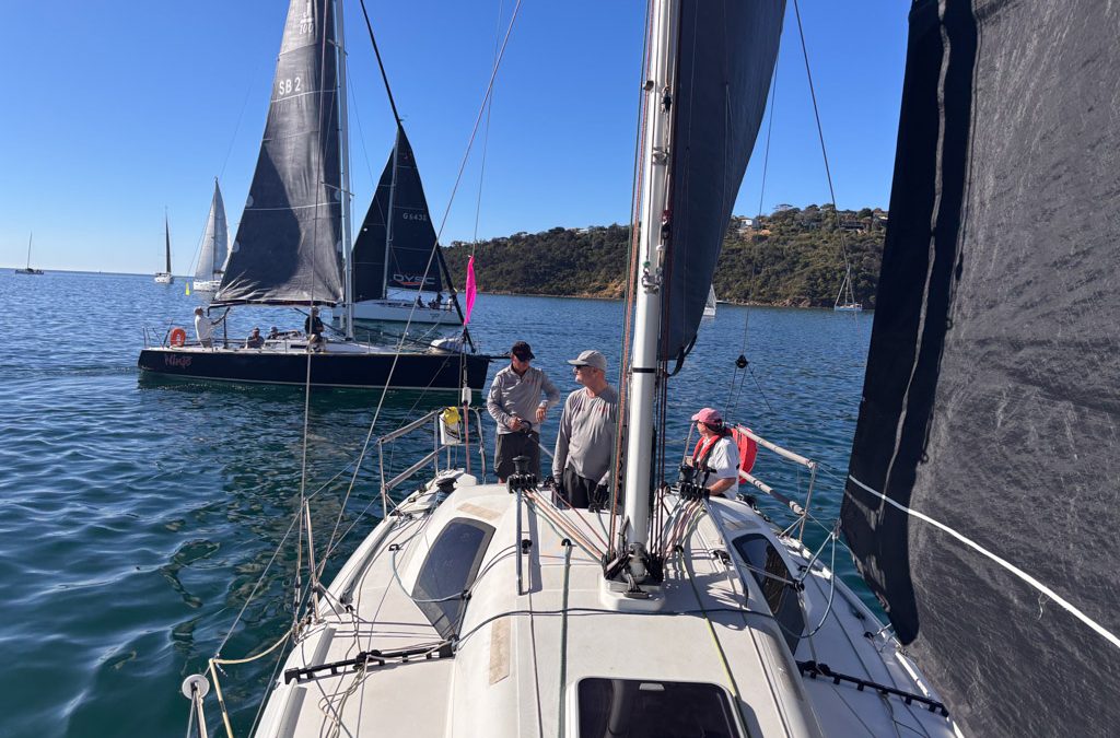 Shaun McKenna on the bow of Unprecedented with jib and mainsail up, other race boats visible in background during very light winds at Sail Peninsula Regatta.