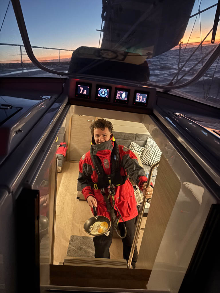 Breakfast on Watch — Frank McKenna in the Galley Looking into the galley of the Hanse 458 where Frank McKenna stands at the bottom of the companionway stairs, holding a saucepan with a freshly cooked omelette. Through the hatch above him, a vibrant orange sunrise fills the skyline, with the four cabin-top B&G instruments beautifully framed and lit in perfect balance.