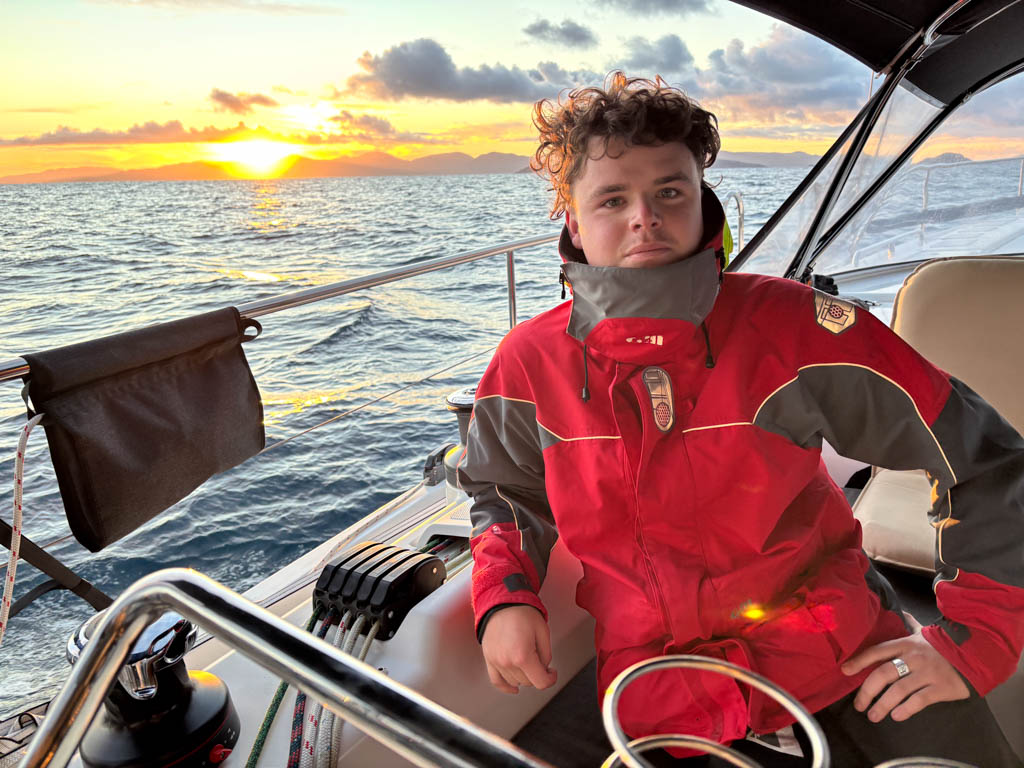 Ocean Seas Yacht Delivery - Offshore Sailing Frank McKenna looking toward the camera at sunrise while on watch during an offshore yacht delivery, dressed in a warm red offshore sailing jacket with the golden morning sun rising over the ocean in the background.