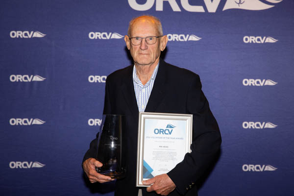 Rik Head standing against the ORCV media wall holding his framed certificate and glass trophy