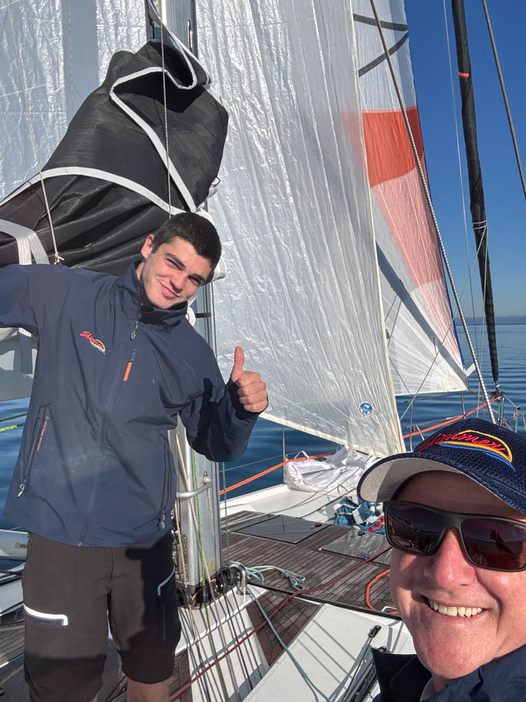 Onboard shot of Sam Backwell giving a thumbs up alongside Shaun McKenna, looking forward from the cockpit of the yacht Shimmer. The bow reveals a tightly furled J0, the flying asymmetrical spinnaker, and the newly rigged North Sails staysail drawing power in the light winter breeze.