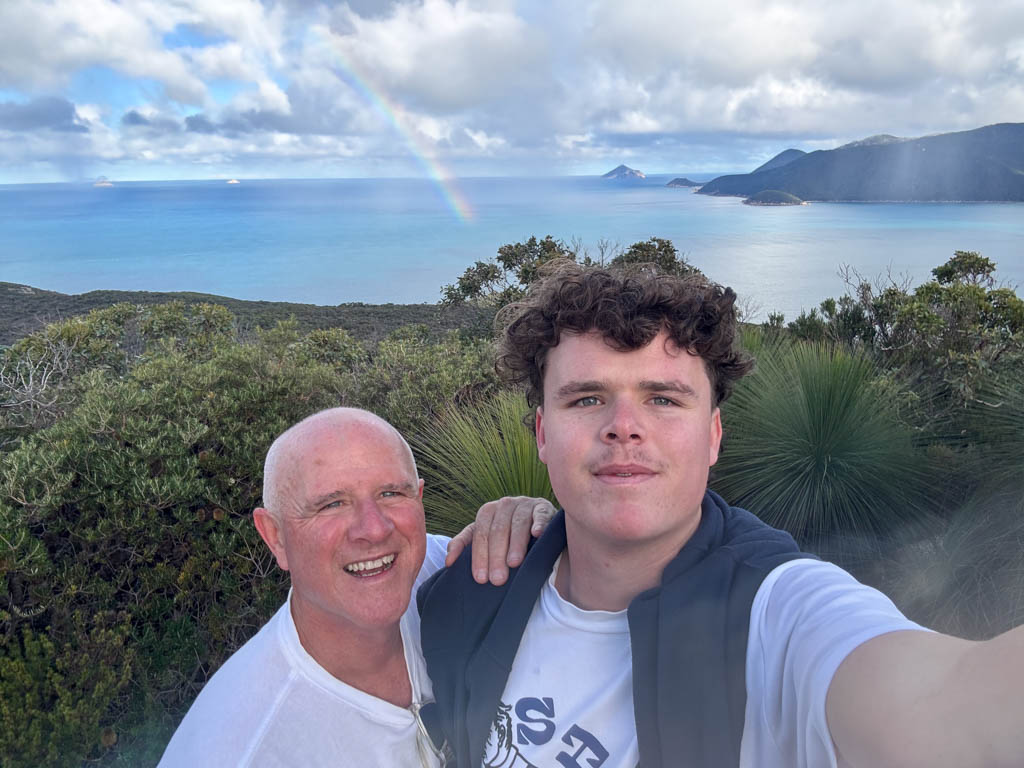 Shaun McKenna and Frank McKenna standing together at the top of the mountain peak at Wilsons Promontory after hiking up from Refuge Cove, with a vast panoramic view of a dead-flat ocean stretching into the horizon behind them.