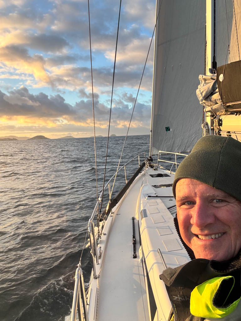 Shaun McKenna on Sunrise Watch Shaun McKenna on deck during an offshore passage at sunrise, with the yacht's jib sail visible against a beautiful orange and blue tinted sky with rolling clouds over a flat, glassy ocean.
